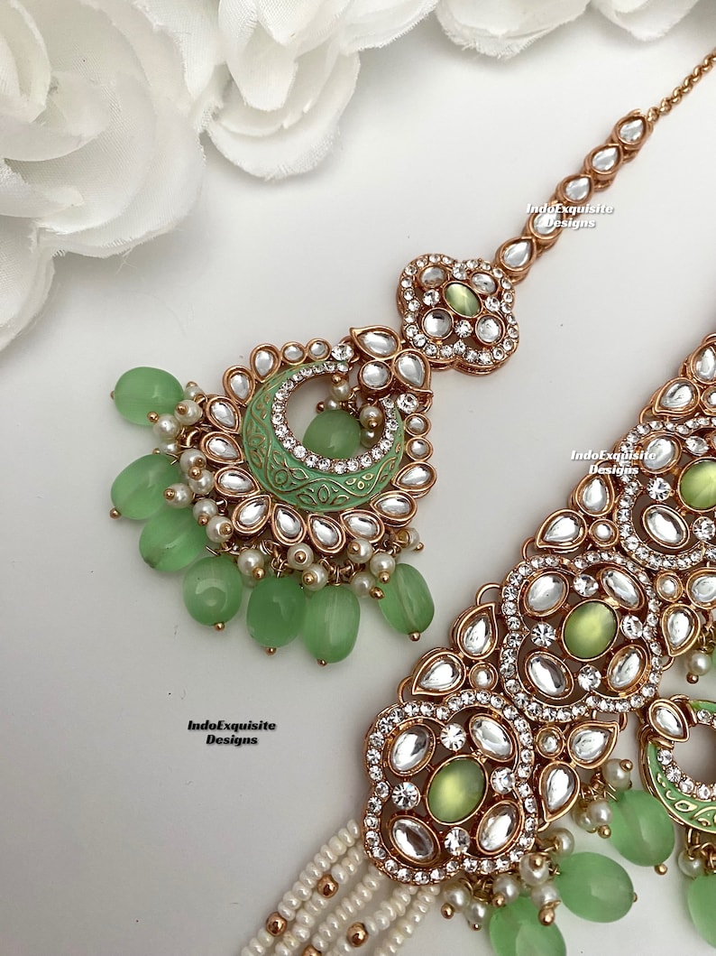 May include: A gold and green Indian bridal headpiece with a large, ornate pendant and a chain of smaller pendants. The pendant features a green stone and is surrounded by clear rhinestones. The chain is adorned with green beads and pearls.