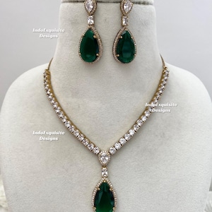 May include: A gold-coloured necklace and earring set. The necklace and earrings feature teardrop-shaped, emerald-green gemstones. The necklace has a row of clear, sparkling stones. The earrings have a clear, rectangular stone above the green teardrop.