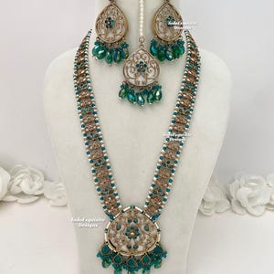 Puede incluir: Un collar de oro y blanco con un gran colgante y cuentas verdes. El collar está sobre un fondo blanco. IndoExquisite Designs está escrito en la imagen.