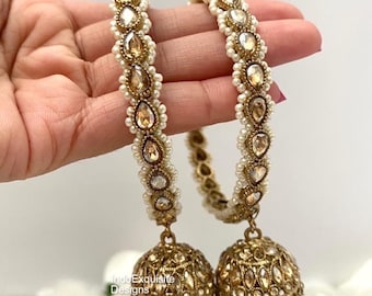 May include: A pair of gold bangles with pearl and clear rhinestone embellishments. Each bangle has a dangling gold jhumka earring with pearl accents.