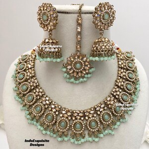 May include: A gold and green Indian bridal necklace set with a matching pair of earrings and a tikka. The necklace features a delicate chain with a pendant, and the earrings are adorned with round stones.