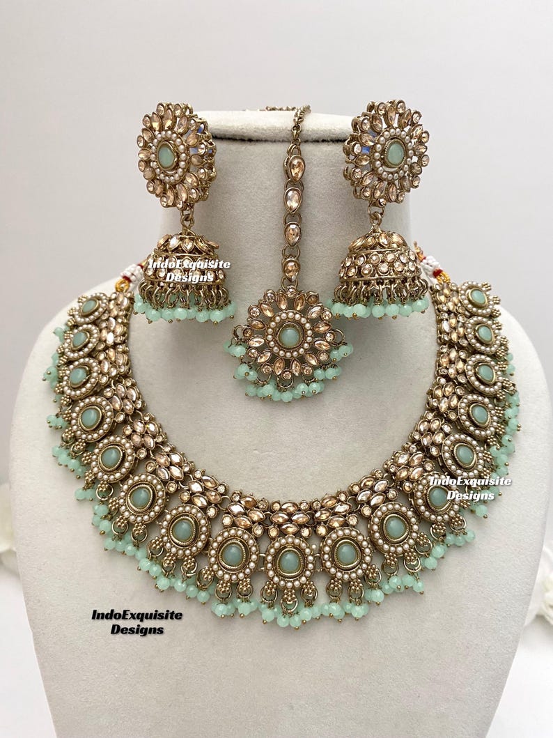 May include: A gold and green Indian bridal necklace set with a matching pair of earrings and a tikka. The necklace features a delicate chain with a pendant, and the earrings are adorned with round stones.