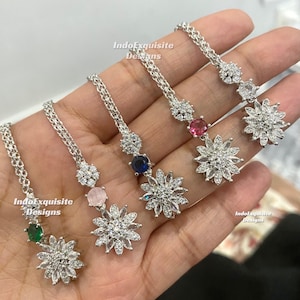 Op de afbeelding: Zilverkleurige sieraden met bloemen- en edelsteenontwerpen. Elk stuk heeft een ketting, een kleine edelsteen en een grotere bloemvormige hanger met heldere stenen. De sieraden worden op een hand getoond.