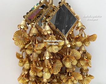May include: Three gold tassel ornaments with a mirror in the centre of each. The tassels are made of gold fabric and have a layered, fan-like design. The mirrors are surrounded by gold sequins. The text "IndoExquisite Designs" is visible on the image.