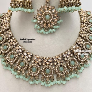 May include: A gold necklace with a delicate design featuring round, light blue stones and pearl accents. The necklace is paired with matching earrings and a maang tikka, a traditional Indian headpiece.