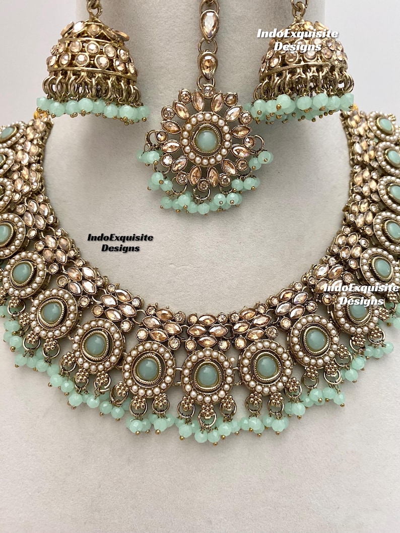 May include: A gold necklace with a delicate design featuring round, light blue stones and pearl accents. The necklace is paired with matching earrings and a maang tikka, a traditional Indian headpiece.