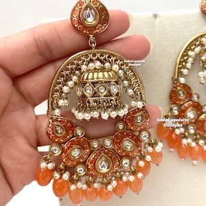 May include: A pair of large, gold-toned earrings with a teardrop-shaped design. The earrings feature a peach-coloured enamel with intricate details and white pearl accents. The earrings are adorned with clear rhinestones and have a dangling bell charm.