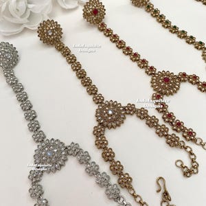 May include: Three gold and silver hand chains with floral designs and jeweled accents. The chains are connected to a ring that can be worn on the finger. The chains are adorned with red, green, and white stones. The text "IndoExquisite Designs" appears on the image.