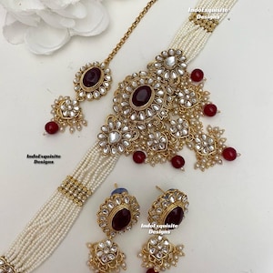May include: A gold necklace with a multi-layered pearl chain and a large, ornate pendant featuring red stones. The pendant is surrounded by smaller pearls and stones. The necklace also includes a matching pair of earrings with red stones and pearl accents. The text "IndoExquisite Designs" appears on the image.