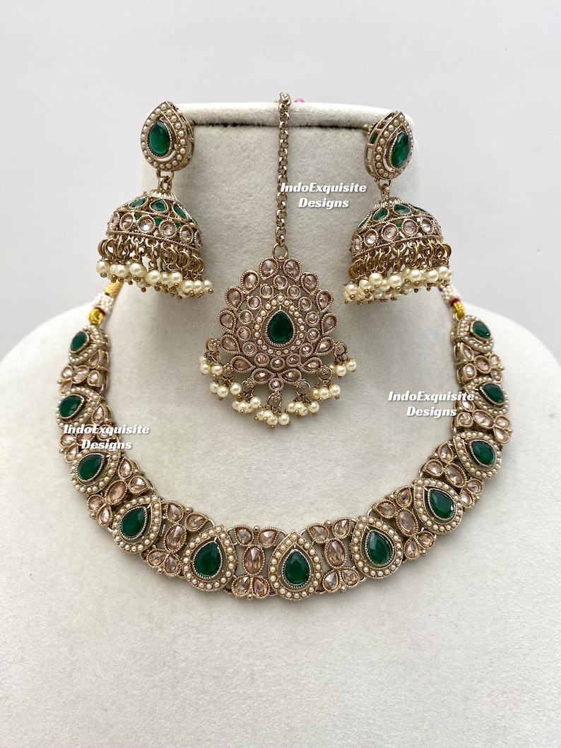 May include: An antique gold-toned jewelry set featuring a necklace, earrings, and a headpiece. The set is adorned with emerald green teardrop-shaped stones and small white pearls. The text "IndoExquisite Designs" is visible.