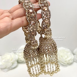 Peut inclure: Une paire de boucles d'oreilles jhumka dorées ornées de pierres claires et pêche. Des perles pendent du bas. Le texte "IndoExquisite Designs" est visible sur l'image.