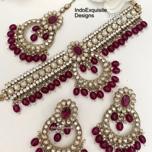 Pode incluir: Um conjunto de joias de noiva indianas tradicionais que inclui um colar choker, um maang tikka e um par de brincos. O conjunto é feito de metal dourado e apresenta designs intrincados com pedras brancas e contas marrons.