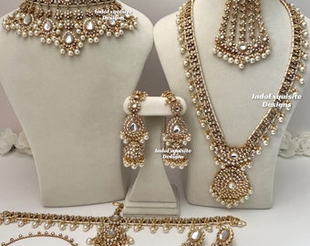 Puede incluir: Un conjunto de joyas de novia indias tradicionales que incluye un collar, pendientes, maang tikka, gargantilla, cinturón y brazaletes de color dorado. El conjunto está adornado con perlas blancas y piedras transparentes.