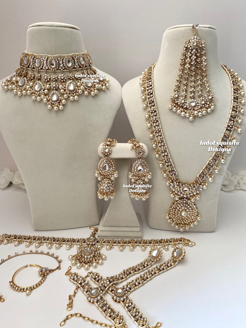 May include: A traditional Indian bridal jewelry set featuring a gold-toned necklace, earrings, a maang tikka, a choker, a waistband, and bangles. The set is adorned with white pearls and clear stones.