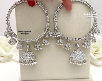 May include: A pair of silver hoop earrings with dangling jhumka charms. The earrings are adorned with clear rhinestones and small pearl accents. The earrings are labeled "IndoExquisite Designs".