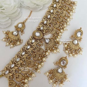May include: A gold-toned jewellery set featuring a wide choker necklace, two drop earrings, and a maang tikka. Each piece displays intricate metalwork, inset white stones, and dangling cream-coloured pearls. The text IndoExquisite Designs is visible.