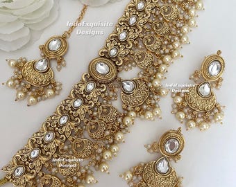 Puede incluir: Un conjunto de joyería en tono dorado que incluye un collar tipo gargantilla ancho, dos pendientes colgantes y un maang tikka. Cada pieza muestra un trabajo de metal intrincado, piedras blancas incrustadas y perlas color crema. El texto IndoExquisite Designs es visible.