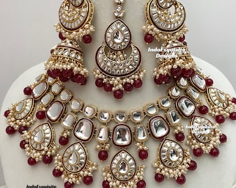 May include: A traditional Indian bridal jewelry set featuring a gold necklace, earrings, and a maang tikka. The set is adorned with intricate designs, clear stones, and red beads.