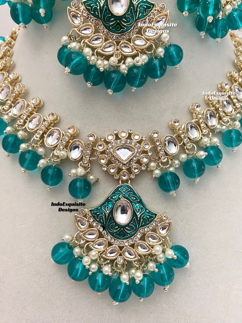 May include: A green and gold necklace with a large pendant featuring intricate designs and clear stones. The necklace is adorned with green glass beads and white pearls.
