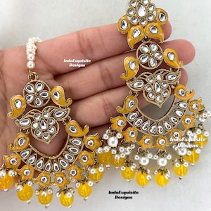 May include: A pair of yellow and gold earrings with white stones and pearl and yellow bead accents. The earrings are hanging from a person's ear and have a floral design. The text "IndoExquisite Designs" is visible on the image.