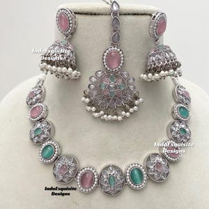 Op de afbeelding: Een zilveren ketting met roze en groene stenen, een bijpassend paar oorbellen en een bijpassende maang tikka. De ketting heeft een bloemmotief en is gemaakt van zilver.