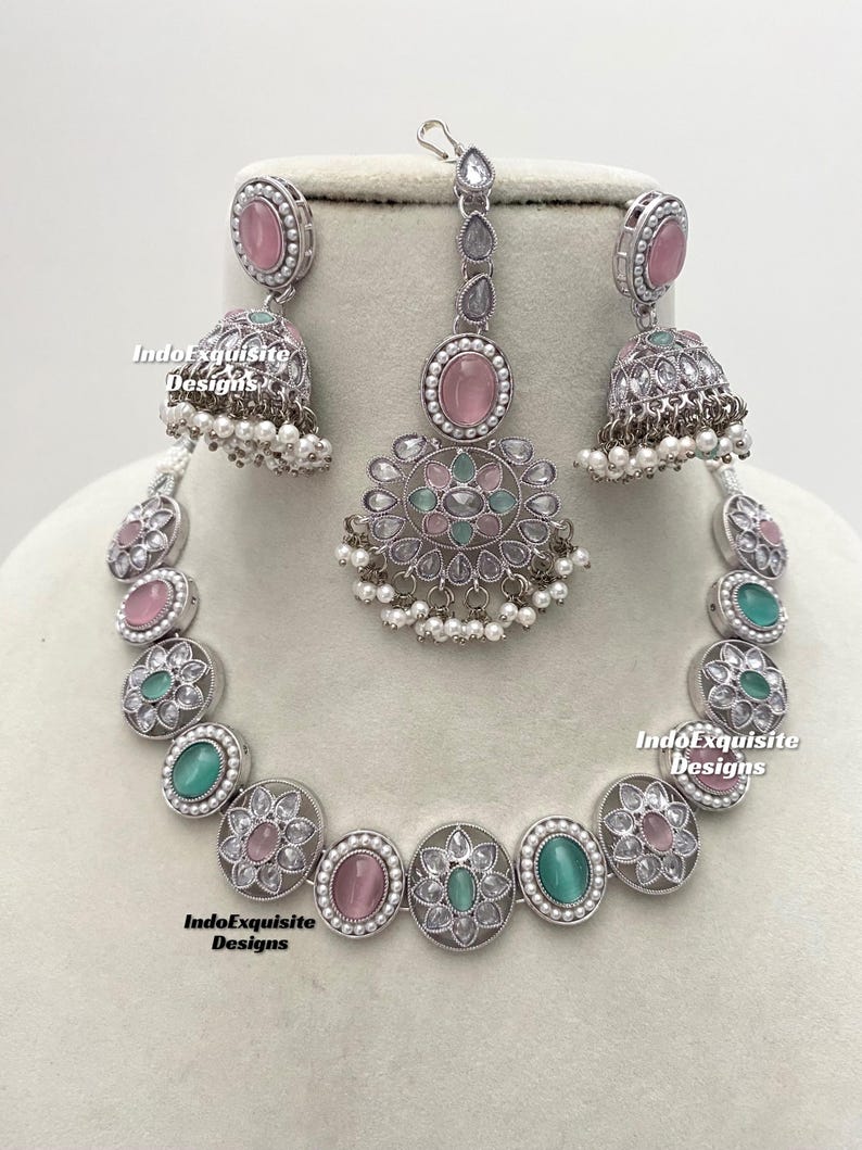 Op de afbeelding: Een zilveren ketting met roze en groene stenen, een bijpassend paar oorbellen en een bijpassende maang tikka. De ketting heeft een bloemmotief en is gemaakt van zilver.