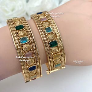 May include: Two gold-colored bangles with colorful rectangular gemstones. The bangles have a textured design and are adorned with green, blue, pink, and yellow stones. The text "IndoExquisite Designs" is visible on the bangles.
