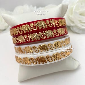 May include: Four bangles, two red and two white, are stacked on a white cushion. Each bangle features a gold elephant design. The text "IndoExquisite Designs" is visible on the bangles.