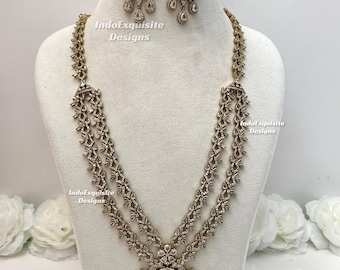 Puede incluir: Un conjunto de collar y pendientes en tono dorado con intrincado trabajo de metal en filigrana, cristales transparentes y piedras azul oscuro en forma de lágrima. El collar tiene un diseño de cadena larga multicapa con un gran colgante central. Texto: IndoExquisite Designs.