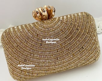 May include: Gold beaded clutch with a decorative floral clasp. The rectangular clutch is covered in small, gold beads arranged in a fan-like pattern. The text "IndoExquisite Designs" is visible on the clutch.