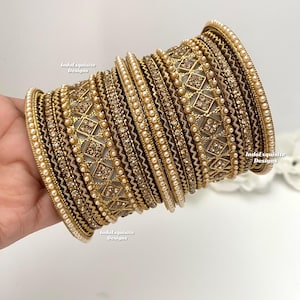 May include: A set of 18 gold bangles with intricate designs and pearl accents. The bangles are stacked on top of each other and have a brown and gold color scheme.