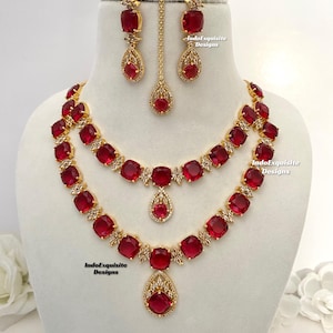以下が含まれることがあります： 赤い金色のネックレスセット。2段重ねで、イヤリングと鼻のリングがセットになっています。ネックレスには大きな赤い石が使用されており、周りを小さな白い石が囲んでいます。イヤリングと鼻のリングにも赤と白の石が飾られています。