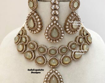 Puede incluir: Un conjunto de joyería que incluye un collar de doble capa, dos pendientes en forma de lágrima y un maang tikka. Cada pieza está elaborada en tono dorado antiguo con piedras cabujón verde claro y cristales. El texto IndoExquisite Designs es visible.