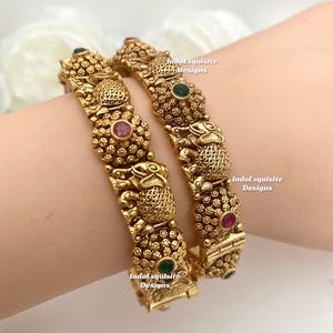 May include: Two gold bangles with detailed patterns and embedded gemstones. The bangles feature intricate designs and are adorned with red and green stones. The text "IndoExquisite Designs" is visible on the bangles.