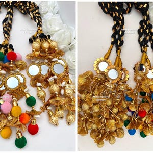 May include: Two ornate gold-toned hair accessories with intricate detailing. Each features a black and gold braided cord, decorative mirrors, and colorful pom-poms in various sizes. The accessories are likely for festive occasions.