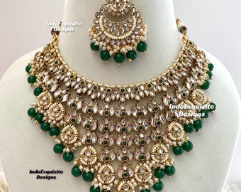 May include: A gold and green Indian bridal necklace set with a teardrop-shaped pendant and a matching maang tikka. The necklace features multiple layers of gold beads and green stones.