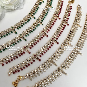 May include: Gold anklets with leaf and pearl designs. The anklets feature clear, oval-shaped stones and small, dangling beads in green, red, and white. The anklets are arranged on a white surface with a floral accent.
