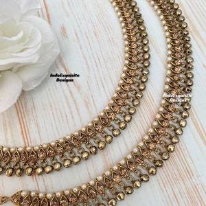 Peut inclure: Une paire de chevillères dorées avec des clochettes pendantes et des accents de cristal. Les chevillères sont ornées de perles blanches.
