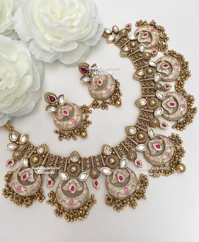 Pu&ograve; includere: Un set di gioielli color oro composto da collana e orecchini. Il set include una collana con pi&ugrave; pendenti a forma di mezzaluna e orecchini abbinati. Ogni pezzo ha una base color crema con motivi floreali rosa e accenti rossi. I gioielli sono impreziositi da cristalli trasparenti.
