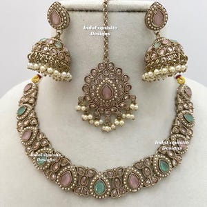 May include: An antique silver-toned jewelry set featuring a necklace, earrings, and a headpiece. The necklace and earrings have pink and teal teardrop-shaped stones. The headpiece is a pendant with a chain. The earrings are bell-shaped with dangling pearls. The text "IndoExquisite Designs" is visible.