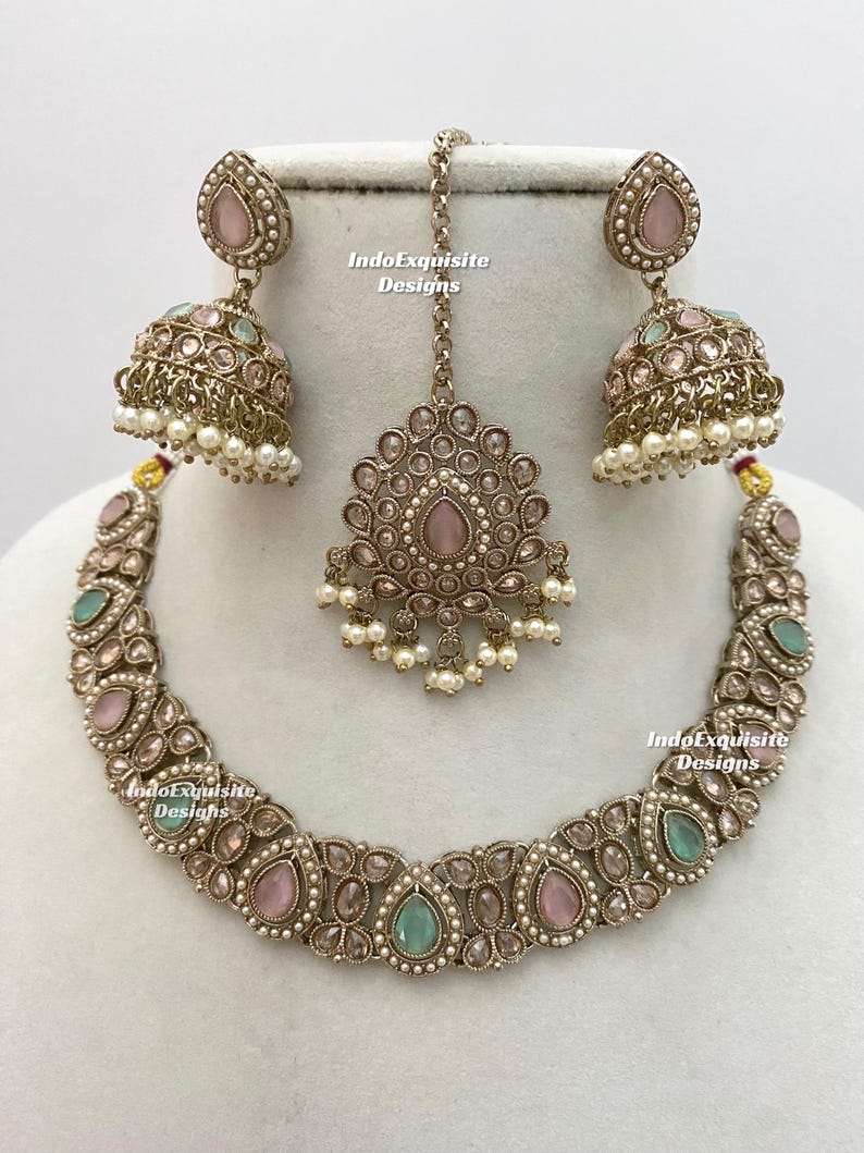 May include: An antique silver-toned jewelry set featuring a necklace, earrings, and a headpiece. The necklace and earrings have pink and teal teardrop-shaped stones. The headpiece is a pendant with a chain. The earrings are bell-shaped with dangling pearls. The text "IndoExquisite Designs" is visible.