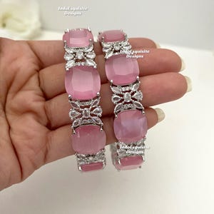 Peut inclure: Une paire de bracelets roses et argentés avec des pierres claires. Les bracelets sont décorés de motifs floraux et le texte "IndoExquisite Designs" apparaît sur chaque bracelet.