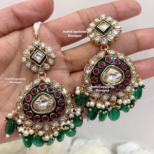 May include: Gold-toned earrings with a square green and white top, and a large, round pendant. The pendant features a central clear stone, surrounded by red stones, white accents, and green and gold detailing. Green and white beads dangle from the bottom.