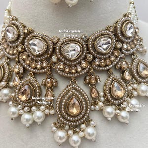 May include: An ornate gold-toned necklace and earring set. The necklace features large, clear, and teardrop-shaped gemstones, surrounded by smaller clear stones and faux pearls. The earrings have a similar design. The set is likely for special occasions.