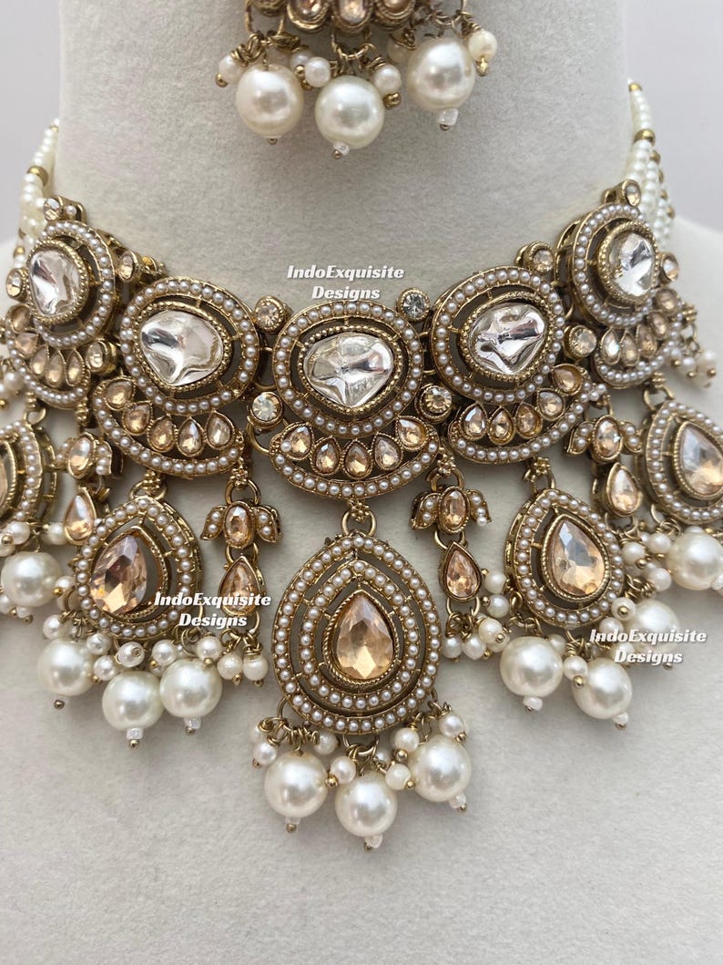 May include: An ornate gold-toned necklace and earring set. The necklace features large, clear, and teardrop-shaped gemstones, surrounded by smaller clear stones and faux pearls. The earrings have a similar design. The set is likely for special occasions.