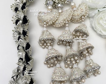 Puede incluir: Un accesorio decorativo colgante con un cordón trenzado negro adornado con flecos metálicos plateados. Incluye varios adornos en forma de cúpula de color crema, decorados con perlas e hilos plateados, con la etiqueta IndoExquisite Designs.