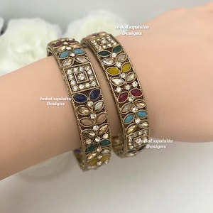 May include: Two gold-coloured bangles with colourful gemstones. One bangle features a square design with a central square of clear stones. The other bangle has a floral design with various coloured stones. The bangles are worn on a wrist.