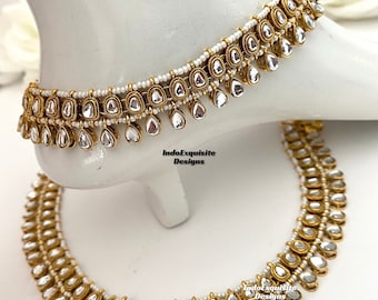 Può includere: Cavigliere dorate con pietre trasparenti a forma di goccia e piccole perline bianche. Le cavigliere sono esposte su un piedino bianco. Il testo "IndoExquisite Designs" è visibile.