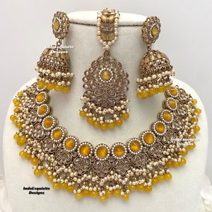 Peut inclure: Un ensemble de collier de mariée indien en or et jaune composé d'un collier ras du cou, de boucles d'oreilles et d'un maang tikka. Le collier présente un grand pendentif rond avec une pierre jaune et des détails complexes. Les boucles d'oreilles sont également rondes et présentent un design similaire. Le maang tikka est une petite pièce délicate avec une pierre jaune.