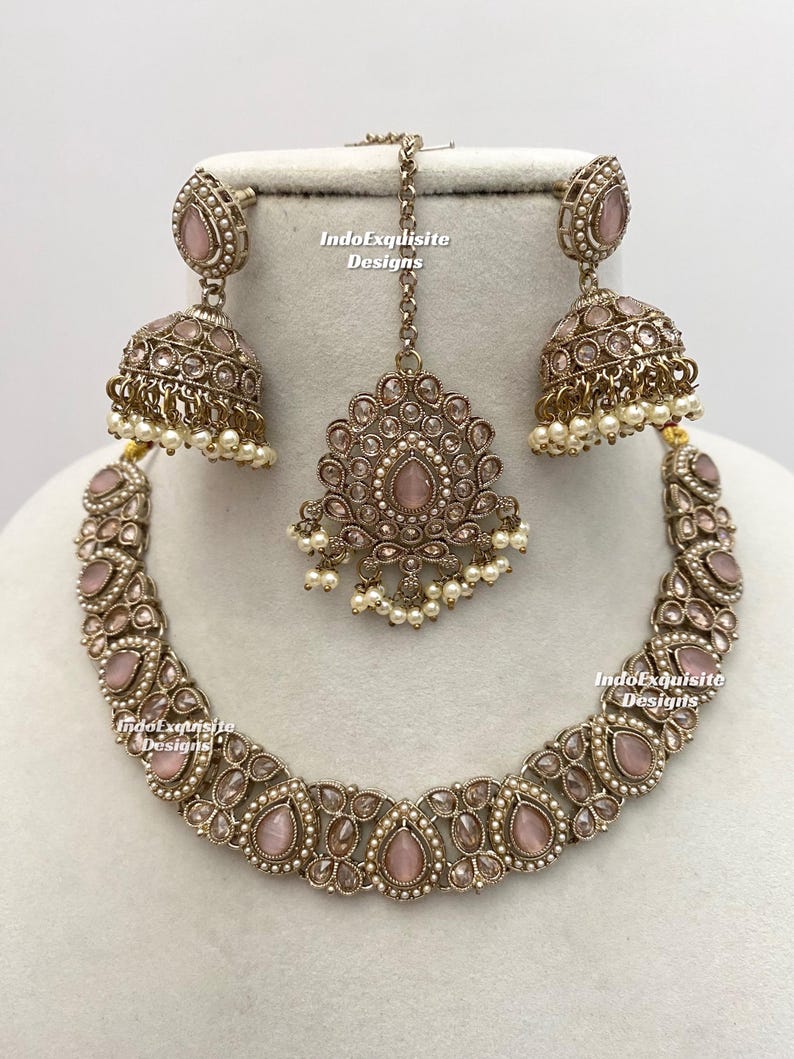 May include: A complete jewelry set featuring a necklace, earrings, and a headpiece. The set is in a silver tone with pink gemstones and small white pearls. The text "IndoExquisite Designs" is visible on the jewelry.